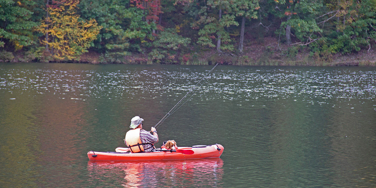 Man wearing a life jacket kayaking with his dog, also wearing a life jacket, and fishing on Poe Lake in Centre County.