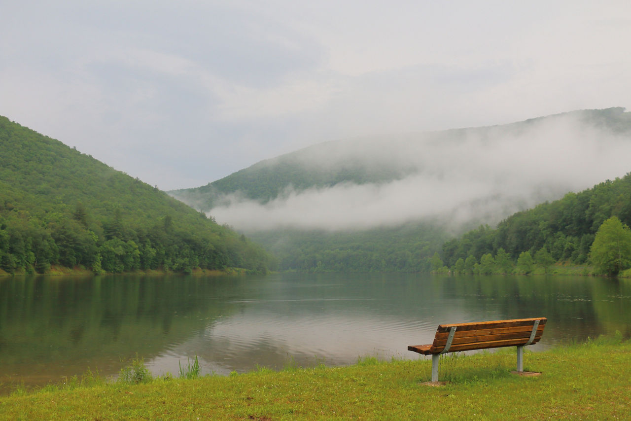 An empty wooden bench along a creek between mountains with fog covering all