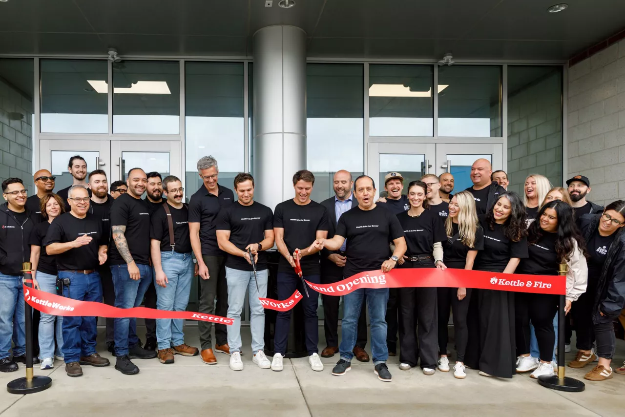 A group of employees wearing shirts with the logo for the company Kettle and Fire cut a ceremonial ribbon outside a new manufacturing building