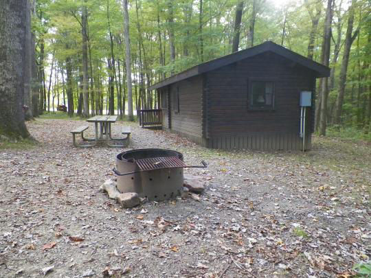 A small wooden building with paved paths to access. The building is surrounded by trees and there is a fire pit out front.