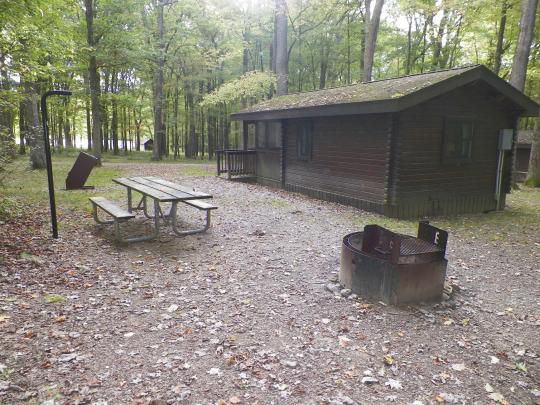 A small wooden building with paved paths to access. The building is surrounded by trees and there is a fire pit out front.