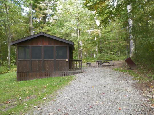 A small wooden building with paved paths to access. The building is surrounded by trees and there is a fire pit out front.