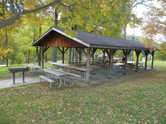 A wooden pavilion covering several picnic tables surrounded by the woods with parking close by