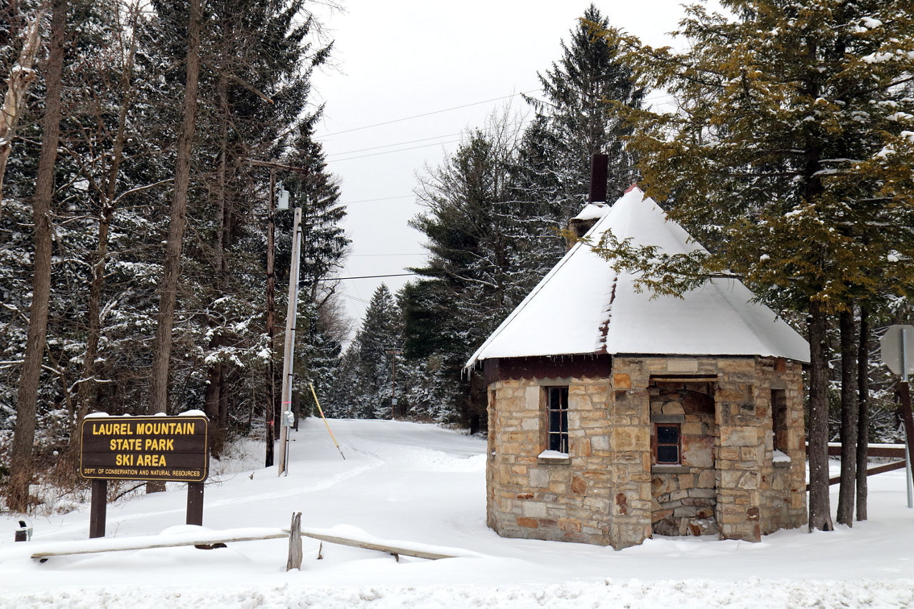 A small, stone, round building next to a sign and evergreens in the background, all covered in a fresh blanket of snow