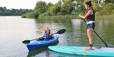 One woman wearing a life jacket kayaking with a registered kayak and another woman wearing a life jacket paddleboarding with a launch permitted paddleboard.