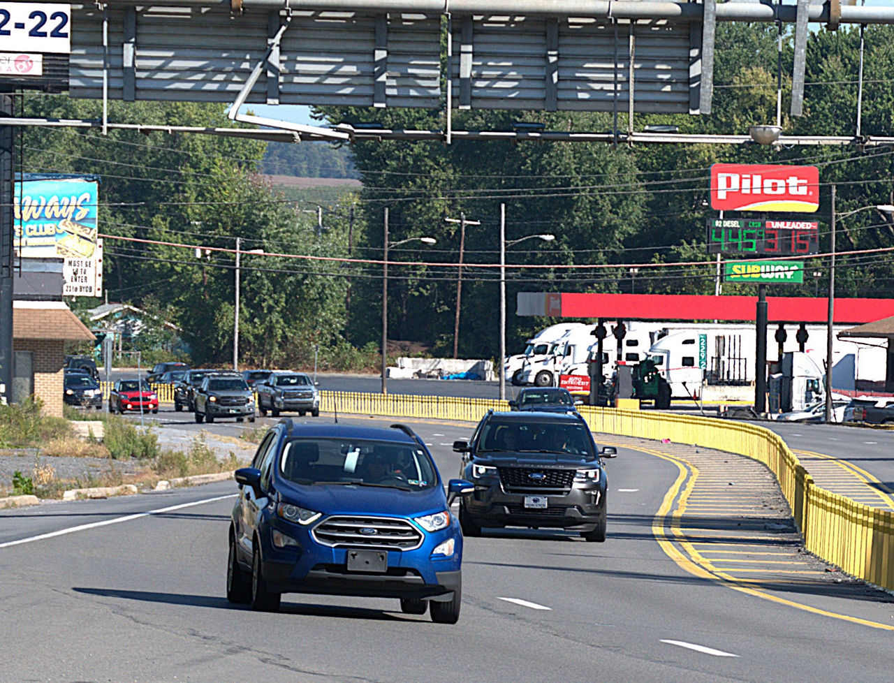 A group of cars drive on a four-lane roadway divided by yellow delineators in the middle of the roadway. A gas station is visible in the background of the photo.