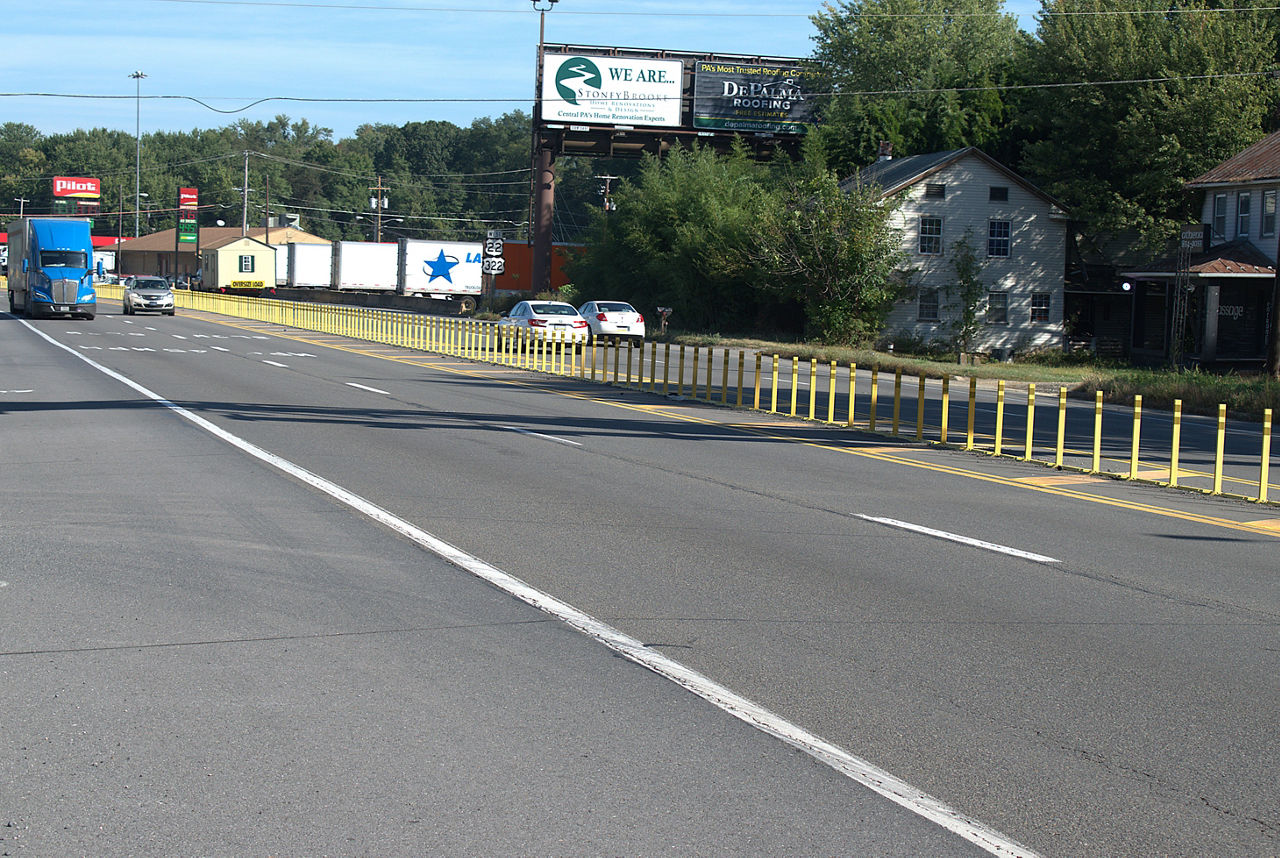 A four-lane roadway divided by yellow delineators in the middle of the roadway with houses, trees, a billboard and gas station on the right side of the roadway. Cars and trucks drive on either side of the roadway.