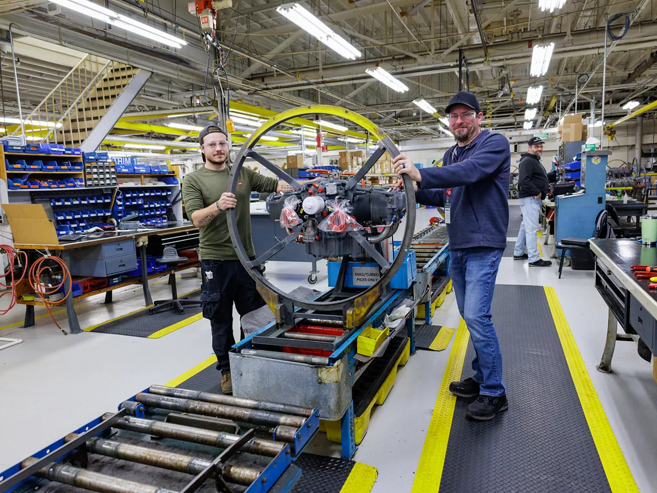 Two manufacturing facility workers pose for a photo while holding a partially-constructed propeller engine.