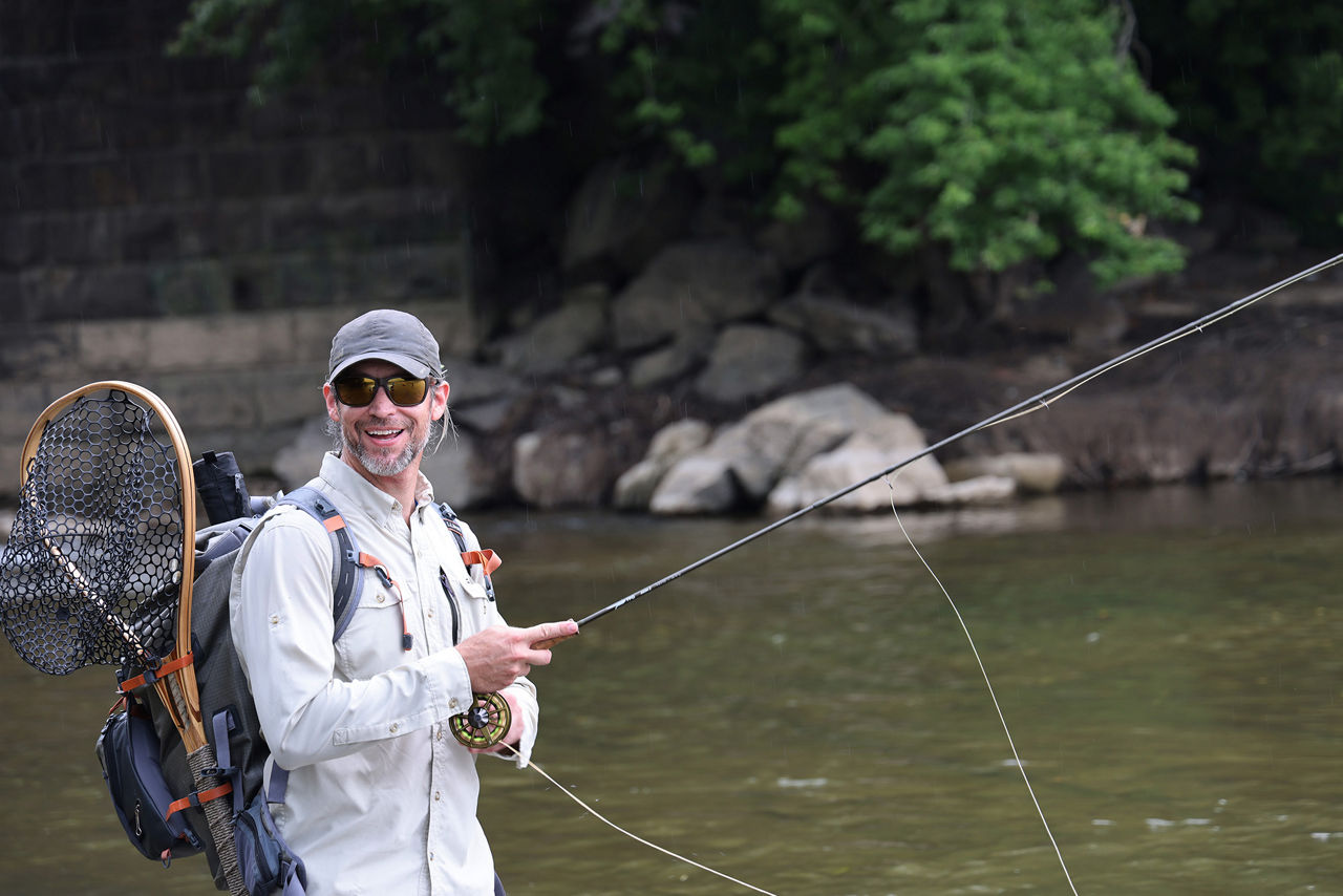 A man wearing a back pack, sunglasses and hat holding a fly fishing rod while standing in a creek.