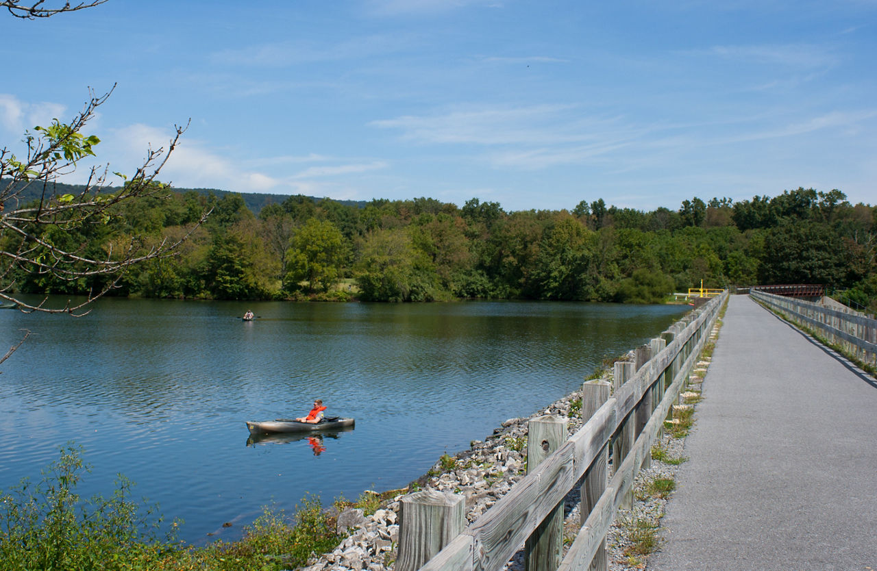 A boater in blue water on a lake with a gravel trail along the dam