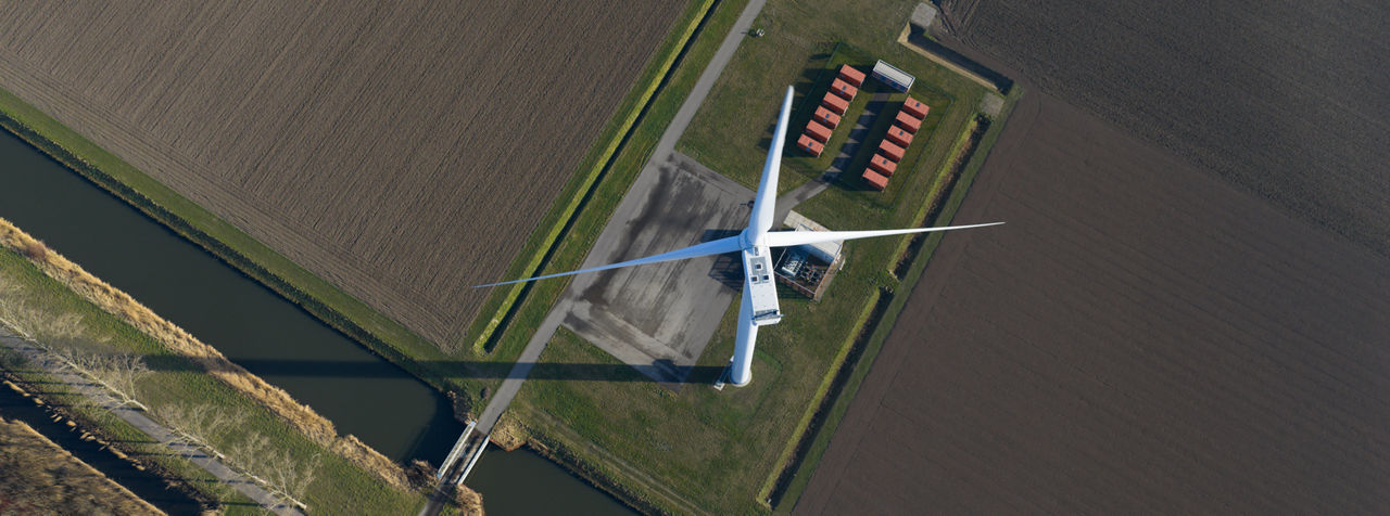 Aerial view of a single wind turbine standing in flat agricultural fields next to a narrow canal.