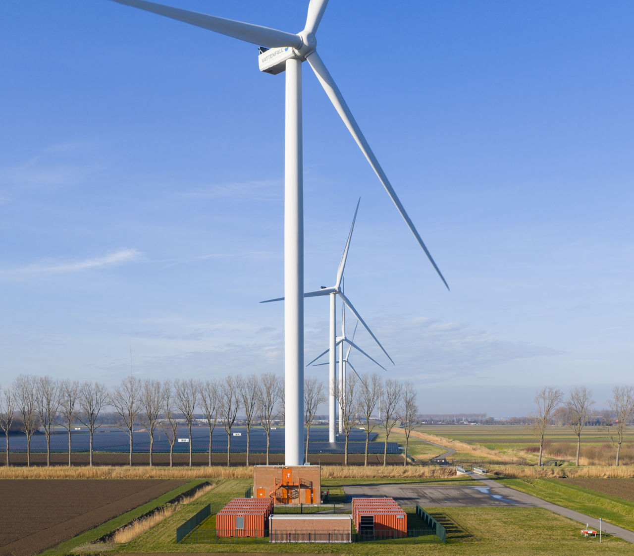 Aerial view of a single wind turbine standing in flat agricultural fields next to a narrow canal.