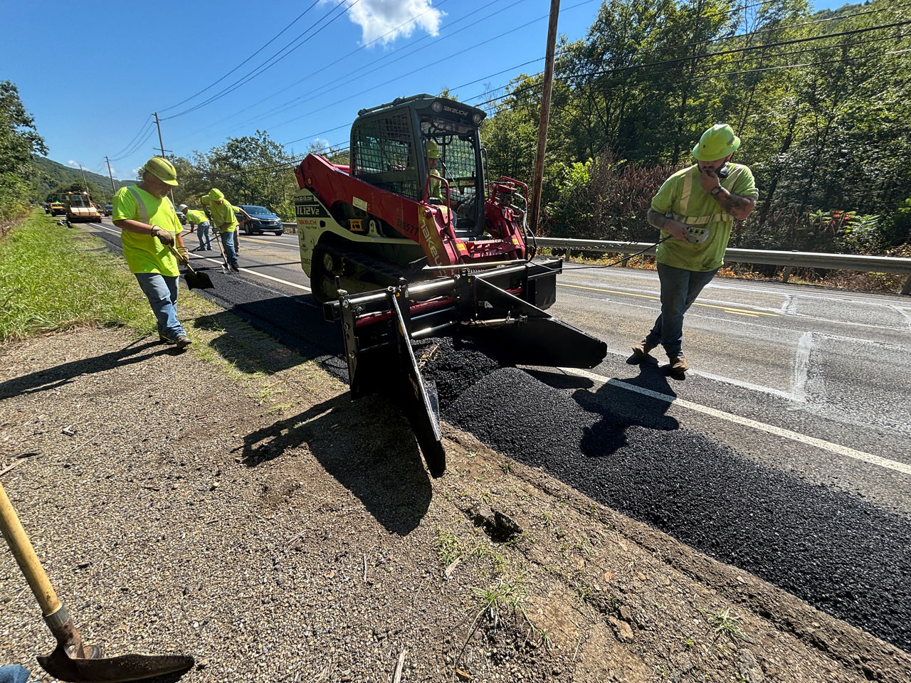  Roadway maintenance crew workers in yellow safety vests and hard hats walk alongside a piece of equipment that is fixing pavement on the edge of a roadway. 