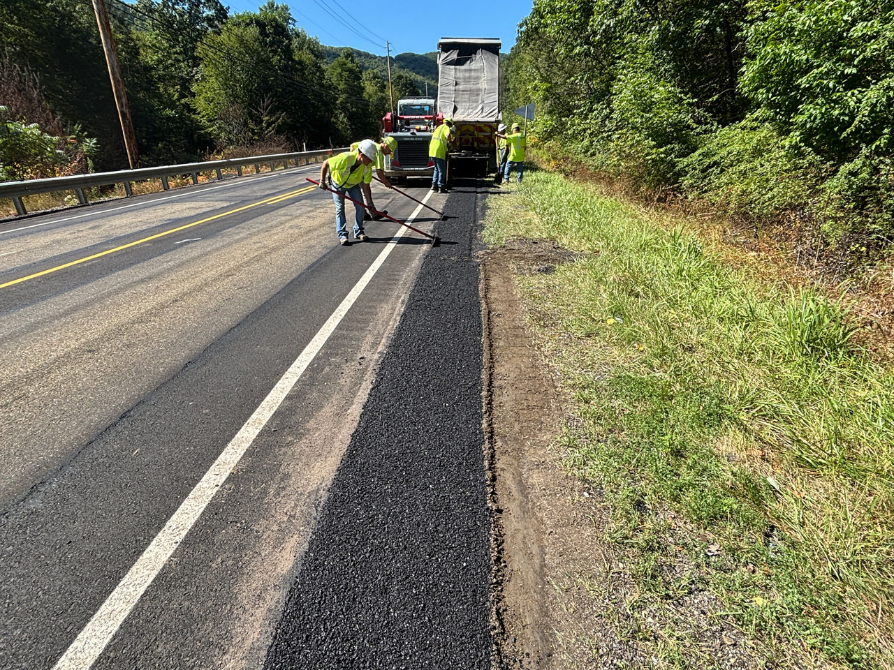 oadway maintenance crew workers in yellow safety vests and hard hats use scrub brooms to help fix pavement on the edge of a roadway. 