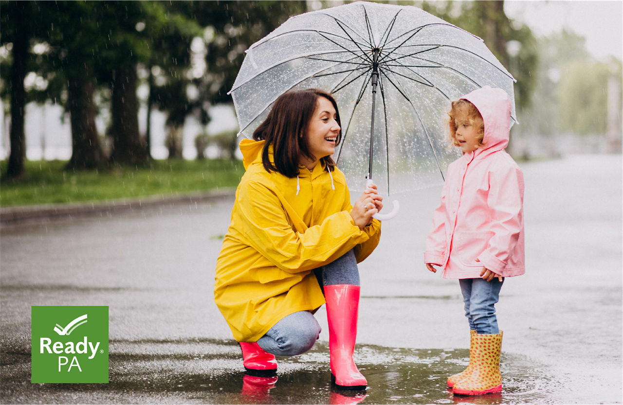 A mom with dark, shoulder-length hair kneels down while wearing a bright yellow rain jacket and bright pink rain boots. She holds a clear umbrella over the head of her small daughter, who is wearing a pink rain jacket with the hood up and patterned rain boots with bright pink soles.