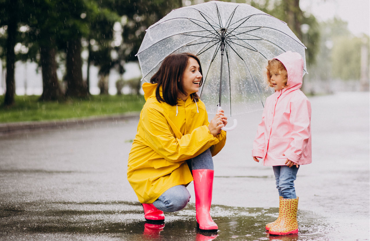 A mom wearing pink boots and yellow raincoat smiles while holding a clear umbrella over a little girl.