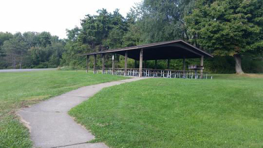 A wooden pavilion covering several picnic tables surrounded by the woods with parking close by