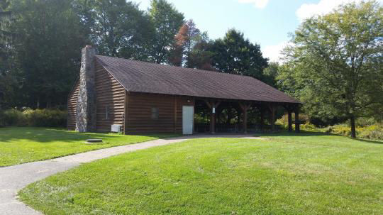 A wooden pavilion covering picnic tables with a stone wall, set in a grassy field, is surrounded by trees
