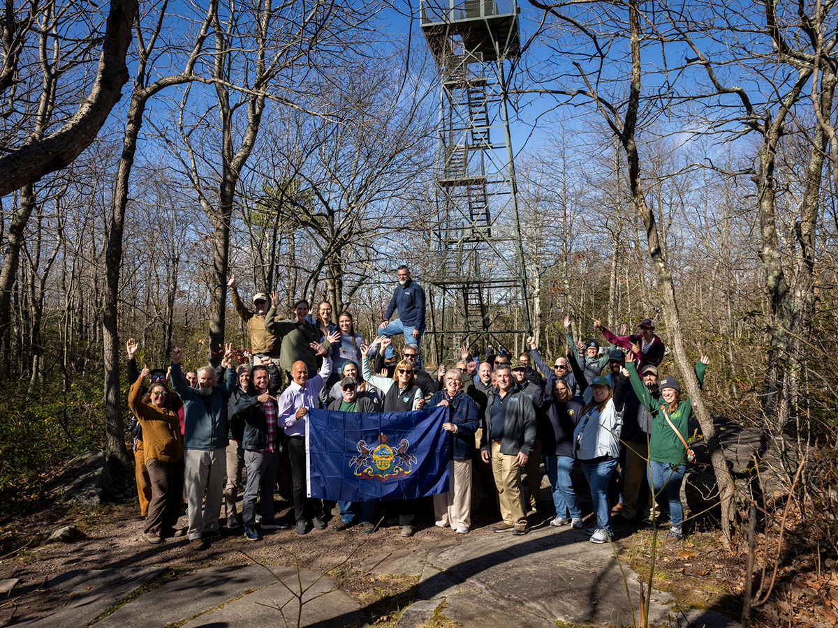 A large group of people posing with a Pennsylvania state flag in front of a tall metal fire lookout tower in a leafless forest on a sunny day.