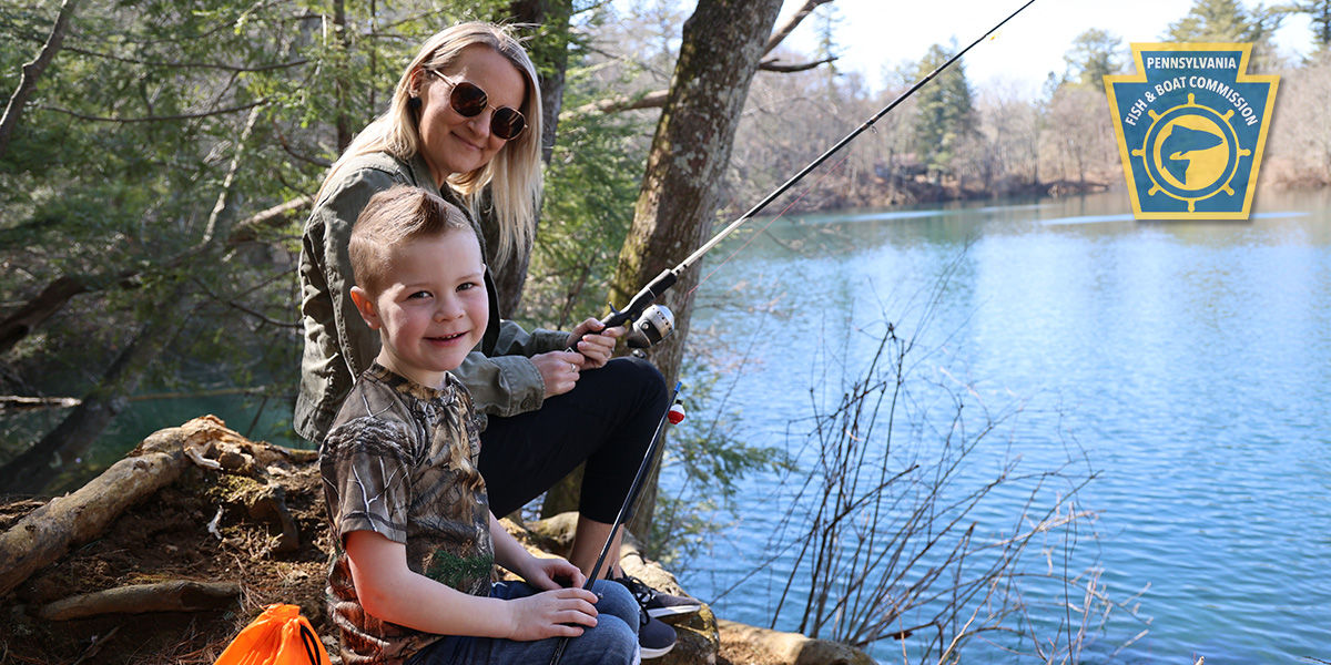 Mother and young son holding fishing rods sit on a forest bank overlooking Fuller Lake.