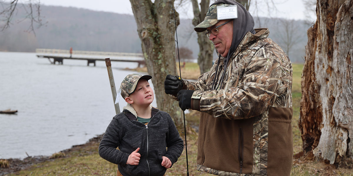 A grandfather rigs a fishing rod for his grandson who is looking up at him admirably. 