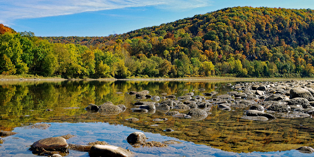 A still and clear river filled with boulders with a fall foliage mountain backdrop