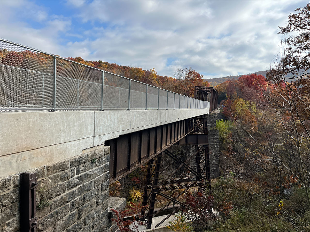 Long steel and concrete bridge crossing a deep valley with autumn-colored trees under a partly cloudy sky.