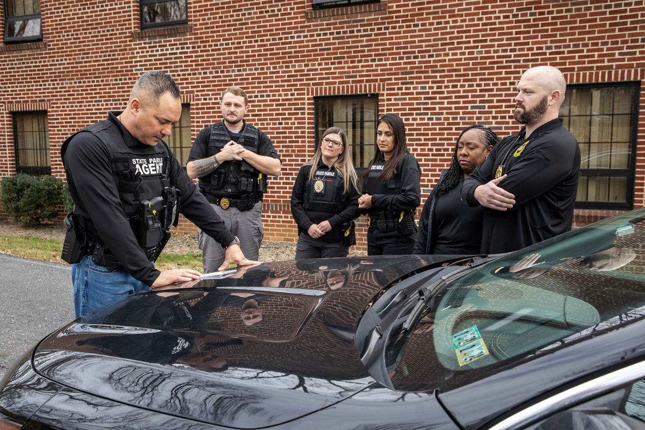 A team of parole agents stand by a car and discuss a mission