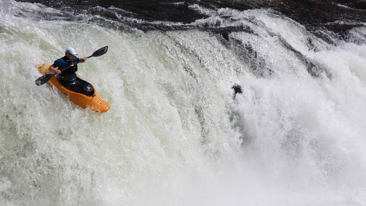 A whitewater kayaker in an orange boat going over a waterfall