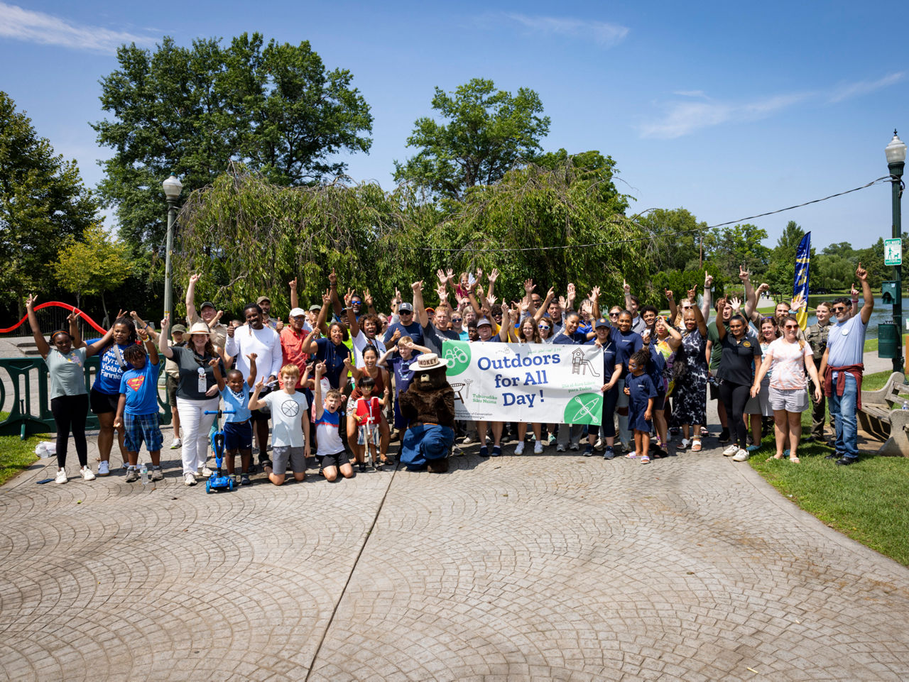 A large group of people pose for a photo in a park while holding an "Outdoors For All Day" event banner.