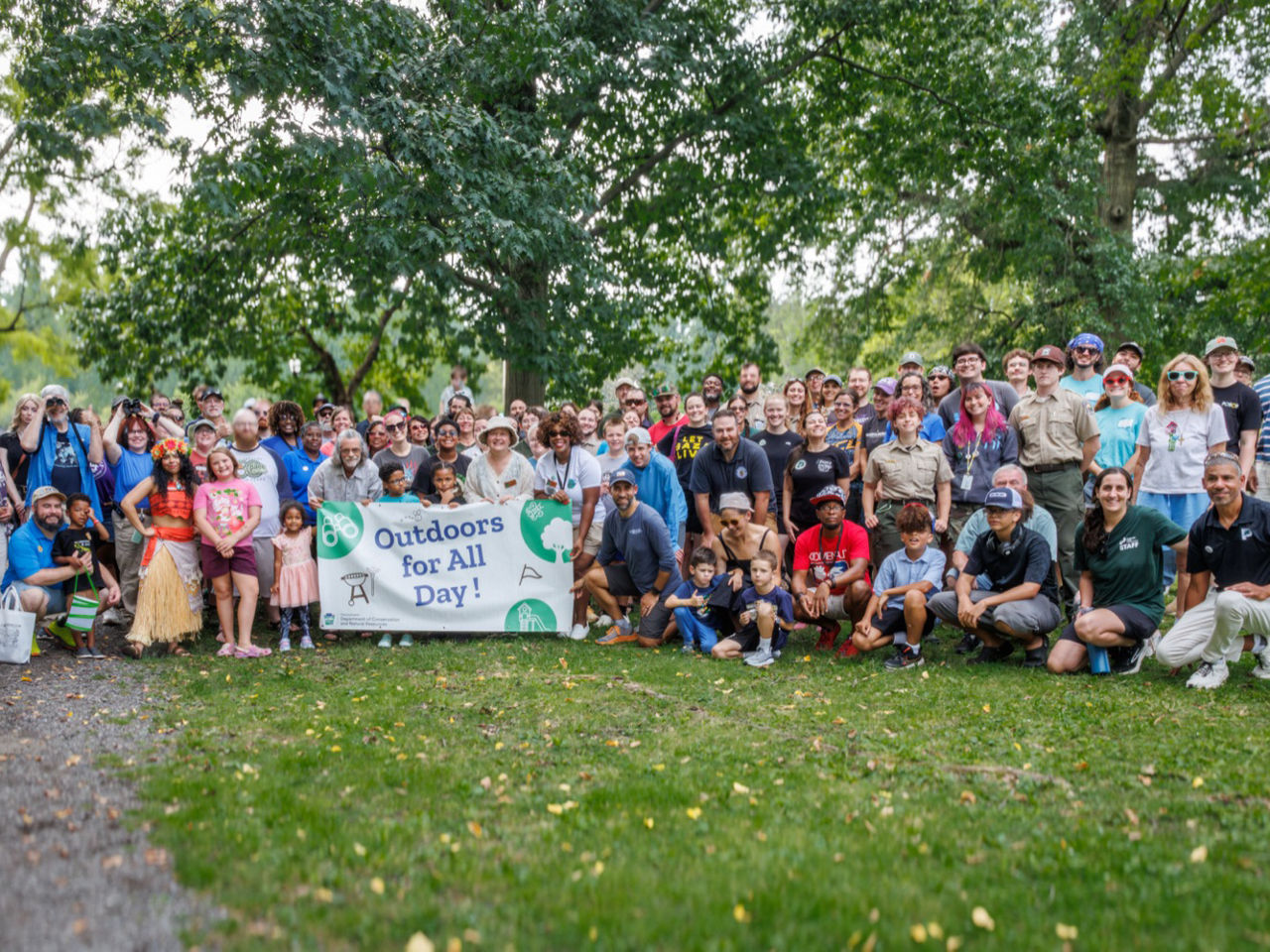 A large, diverse group of people pose for a photo while holding an "Outdoors for All Day" banner.