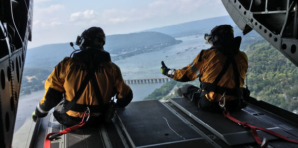 Two members of the Helicopter Aquatic Rescue Team sit in the back of  a helicopter and overlook a river.