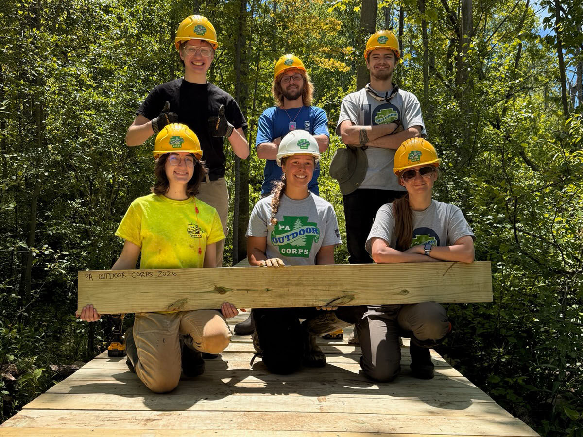 A group of PA Outdoor Corps members hold a wooden plank while standing on a footbridge.