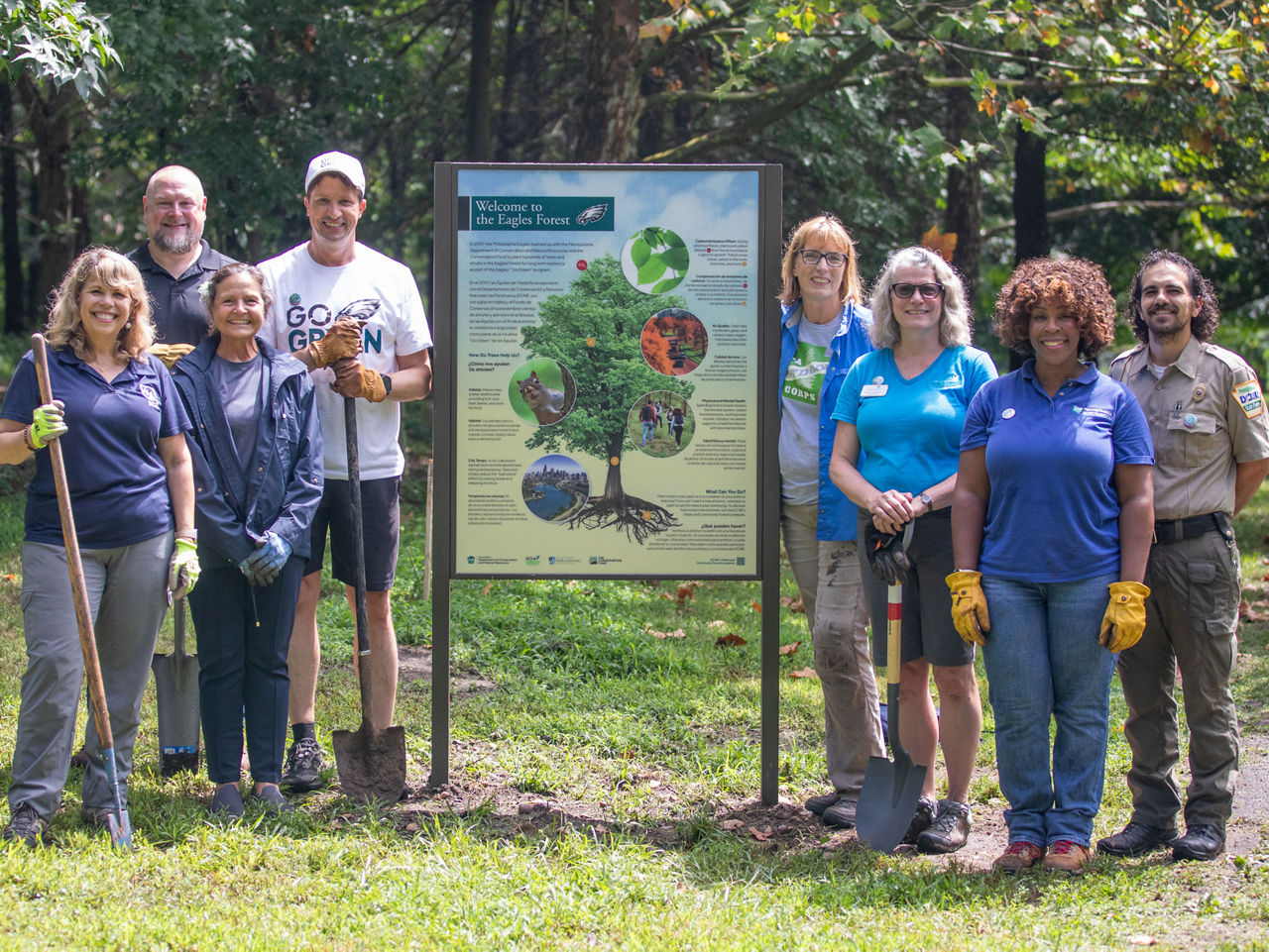 A large group holding various tree planting tools stands around a sign at Eagles Forest in Neshaminy State Park