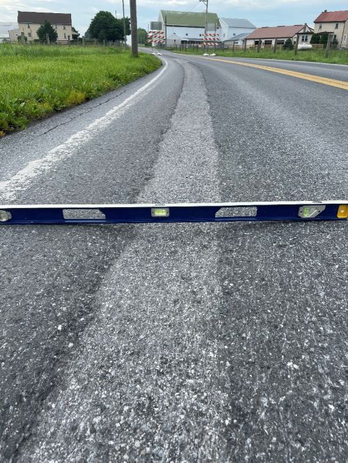 An image of a metal level sitting in the middle of a trough that has developed on the roadway. Grass, trees and houses are in the background of the photo.     