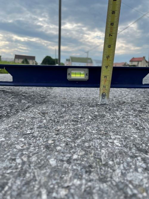 A close-up image of a metal level sitting on a roadway with a measuring tape up against it to show the trough that’s developed on the roadway.  