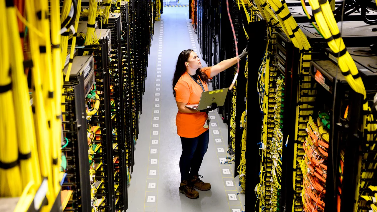 An Amazon employee wearing an orange shirt and safety goggles inspects a row of server racks in a large data center