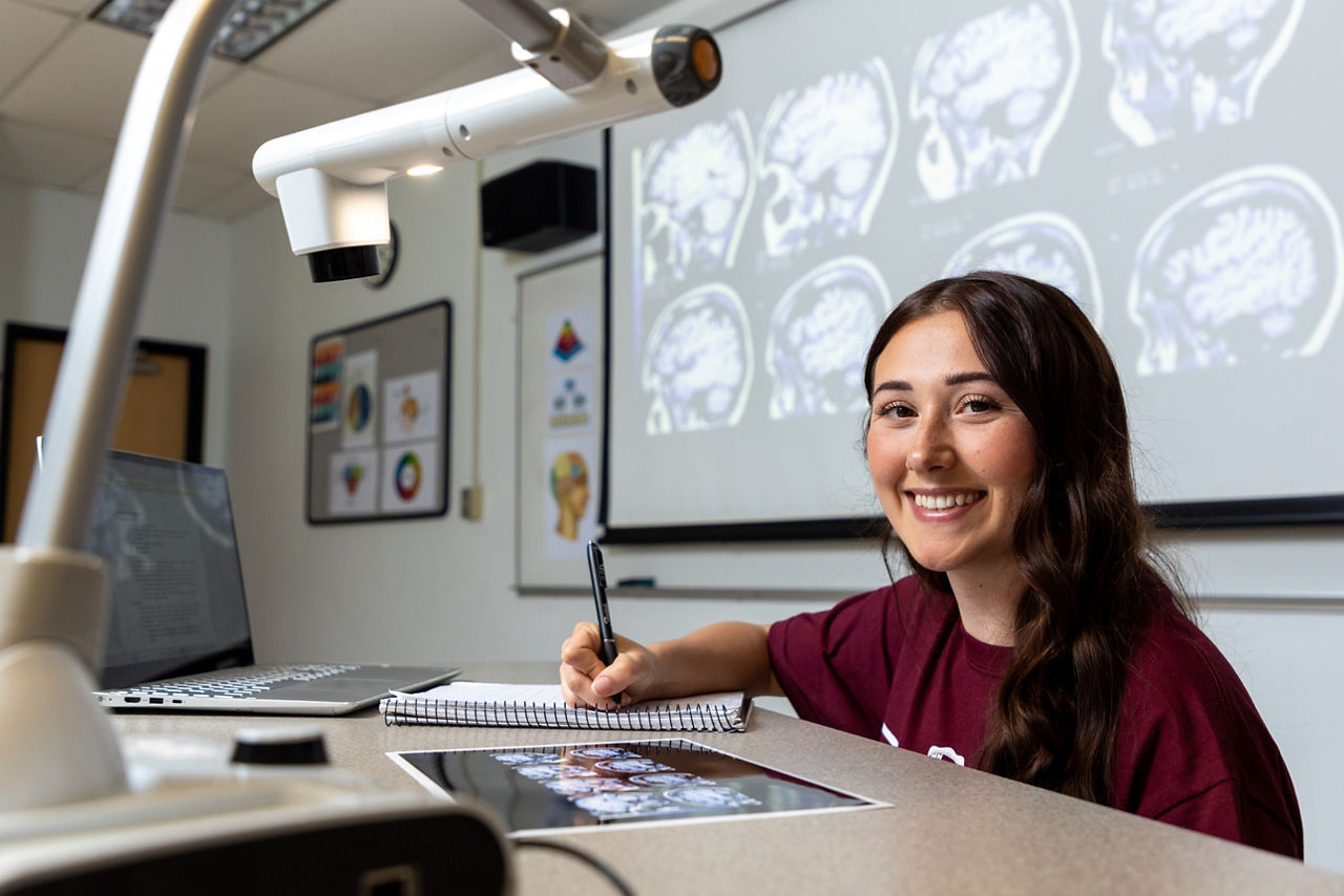 A student at West Chester University, part of Pennsylvania’s State System of Higher Education, smiles while studying brain scans in a classroom. She is seated at a desk with a laptop, notebook, and projector, with MRI brain images displayed on a screen behind her.