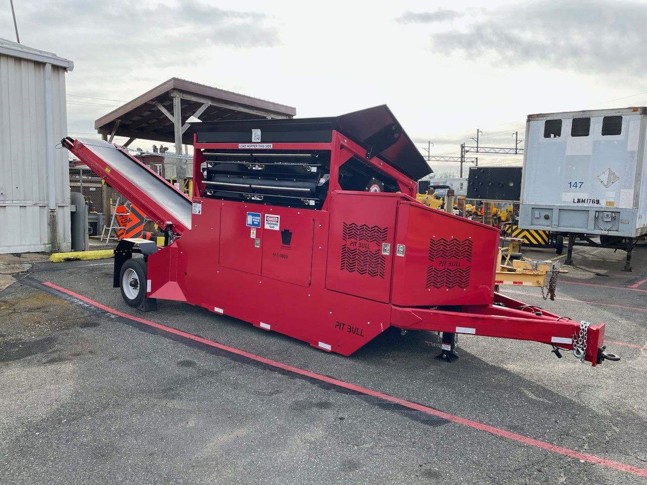 A large red piece of equipment mounted on a trailer chassis with a conveyor belt on one end and the trailer hookup on the other end.