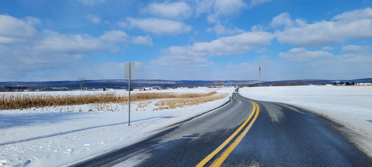 Image of almost dry two-lane roadway with rows of corn functioning as a snow fence of the left side of the roadway.