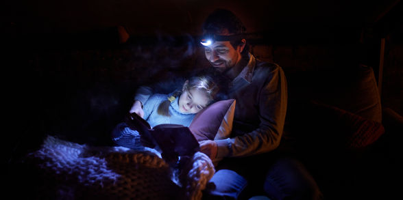 A dad and daughter wear headlamps while reading a book in the dark.