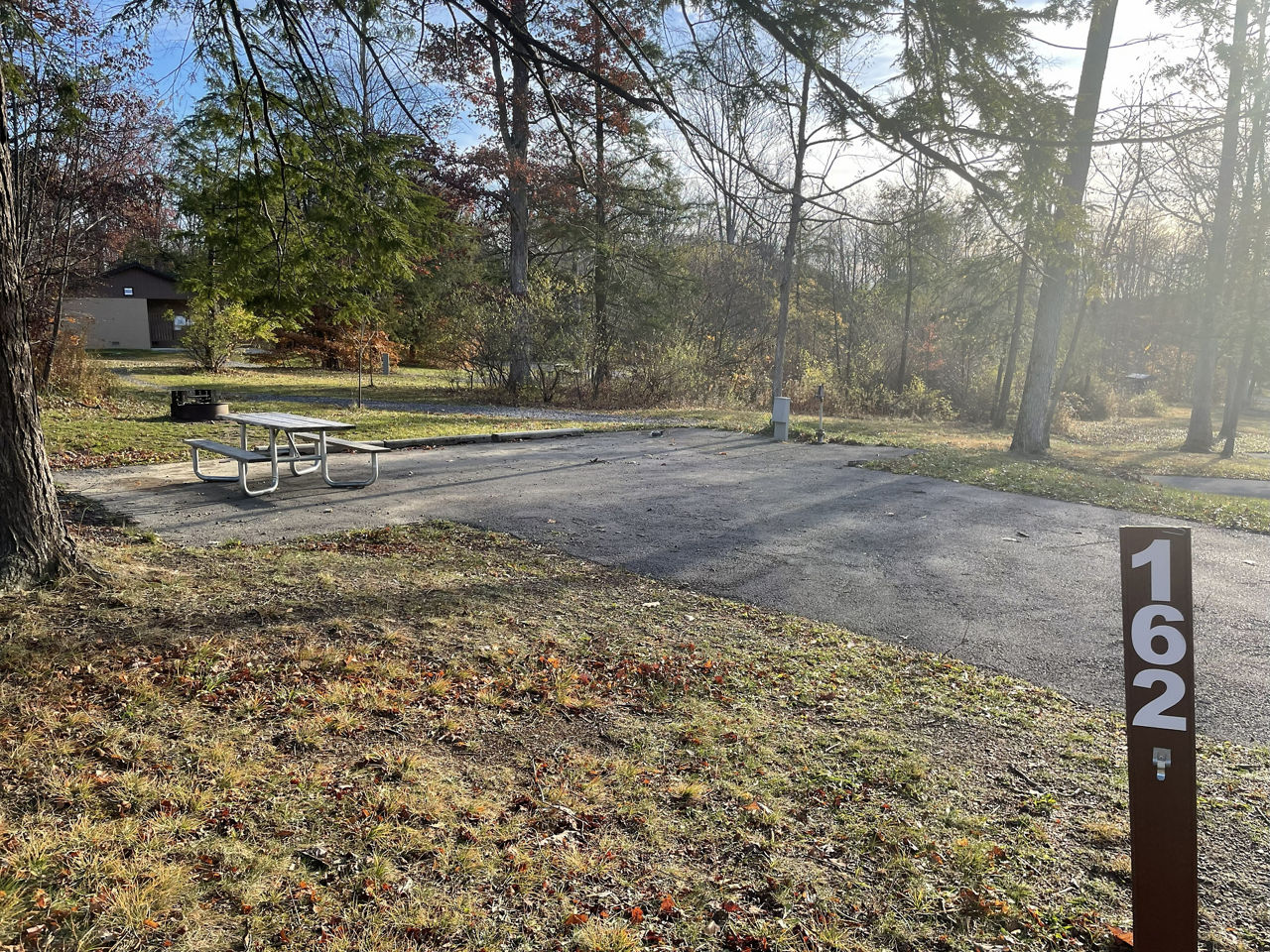 A paved, accessible path meanders through a tranquil forest with bare trees and fallen leaves. A picnic table, designed for easy access, is visible in the background, inviting visitors to enjoy a meal in nature. 