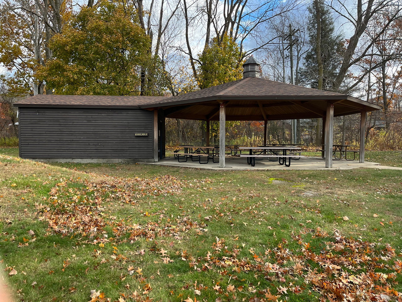 A wooden pavilion covers several picnic tables and is nestled in the woods. An paved path leads to the pavilion.