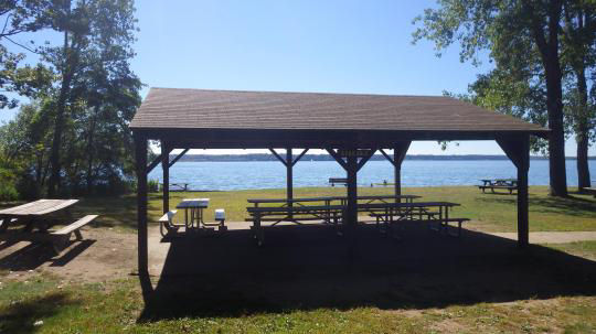 A wooden pavilion covering several picnic tables surrounded by the woods with parking close by