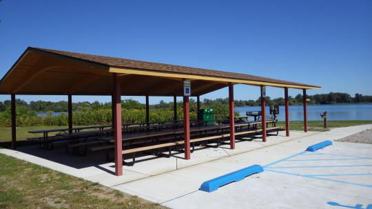 A wooden pavilion covering picnic tables with a stone wall, set in a grassy field, is surrounded by trees
