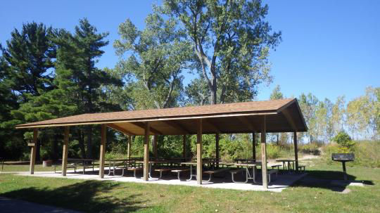 A wooden pavilion covering picnic tables with a stone wall, set in a grassy field, is surrounded by trees