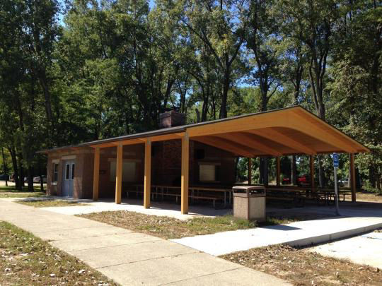 A wooden pavilion covering picnic tables in a grassy field surrounded by trees with a lake in the background.