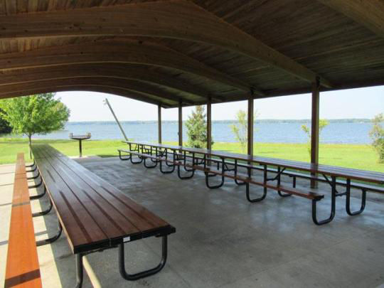 A wooden pavilion covering picnic tables in a grassy field surrounded by trees with a lake in the background.