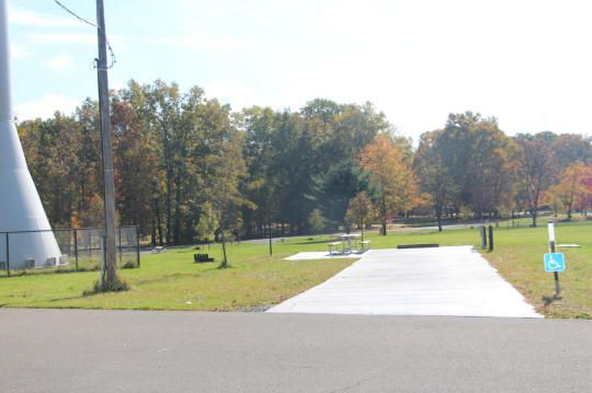 A paved, accessible path meanders through a tranquil forest with bare trees and fallen leaves. A picnic table, designed for easy access, is visible in the background, inviting visitors to enjoy a meal in nature. 