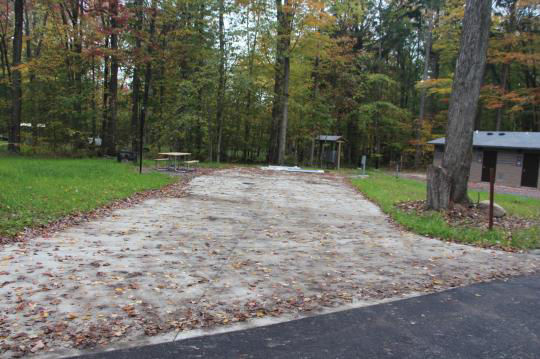 A paved, accessible path meanders through a tranquil forest with bare trees and fallen leaves. A picnic table, designed for easy access, is visible in the background, inviting visitors to enjoy a meal in nature. 
